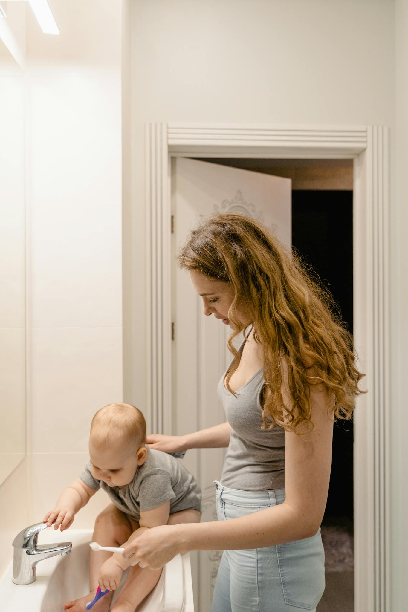 A mother assists her baby in brushing teeth in a bathroom setting.